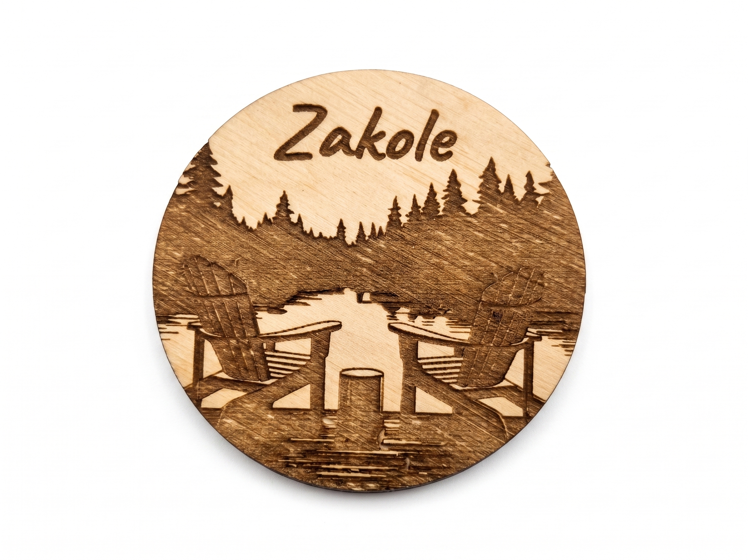 Okrągły magnes z jasnego drewna z wygrawerowanym napisem "Zakole" i sceną leżaków nad brzegiem jeziora otoczonych lasem, na czystym białym tle.