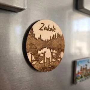 Okrągły magnes z drewna z laserowo wygrawerowanym napisem "Zakole" oraz motywem dwóch leżaków nad wodą wśród lasu, przyklejony do lodówki obok innych magnesów.